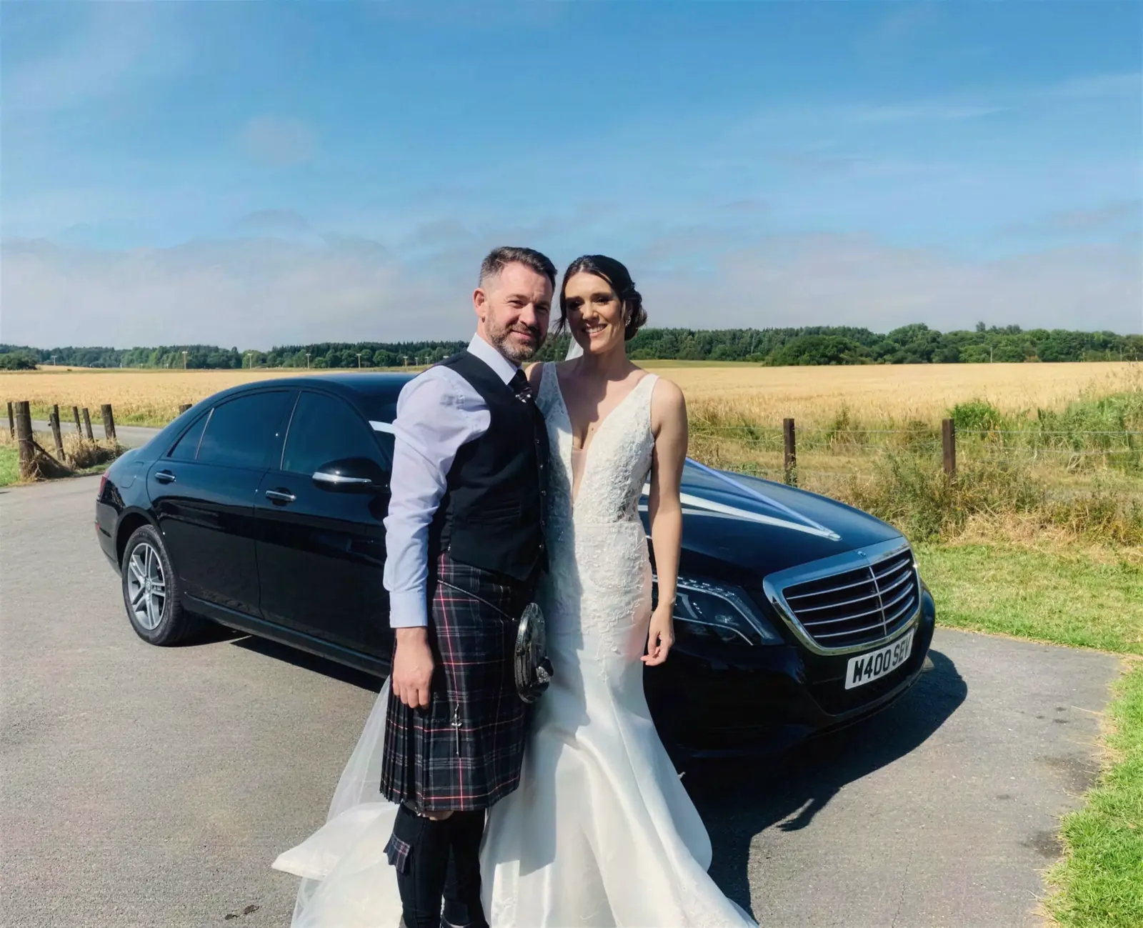 A bride and groom in a kilt beside a black Speyside Executive Hire Mercedes